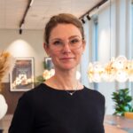 Porträtt av en person med glasögon och mörk tröja som står i en modern inomhusmiljö med stora fönster, växter och dekorativ belysning i bakgrunden.